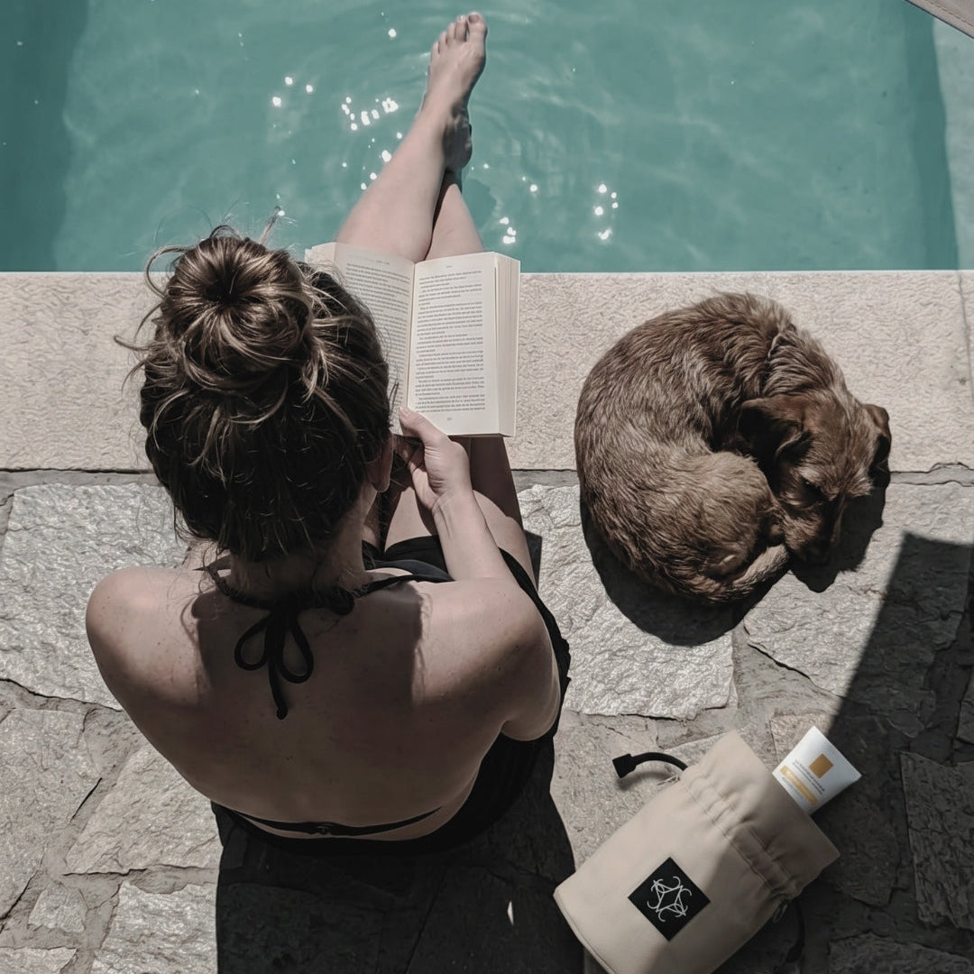 Frau mit Hund am Pool liest ein Buch und sonnt sich.